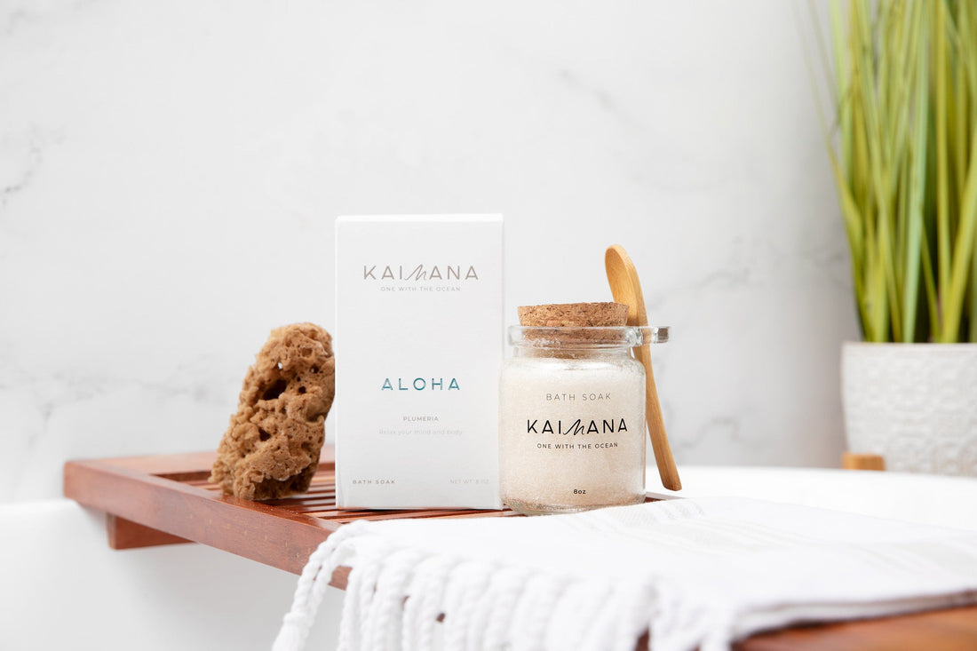 Kaimana Aloha magnesium bath soak jar with cork lid and wooden scoop on a spa bath tray next to product box and natural sponge