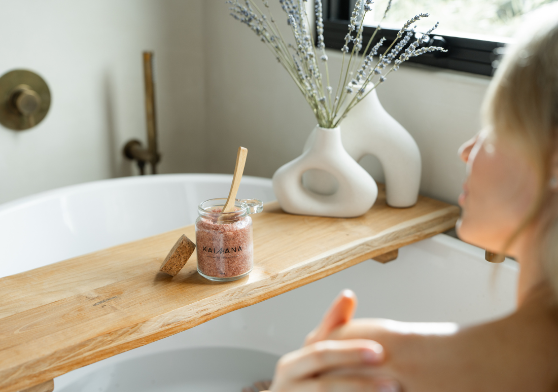 Kaimana mineral bath soak jar with wooden scoop on a bath tray next to a woman relaxing in a bathtub